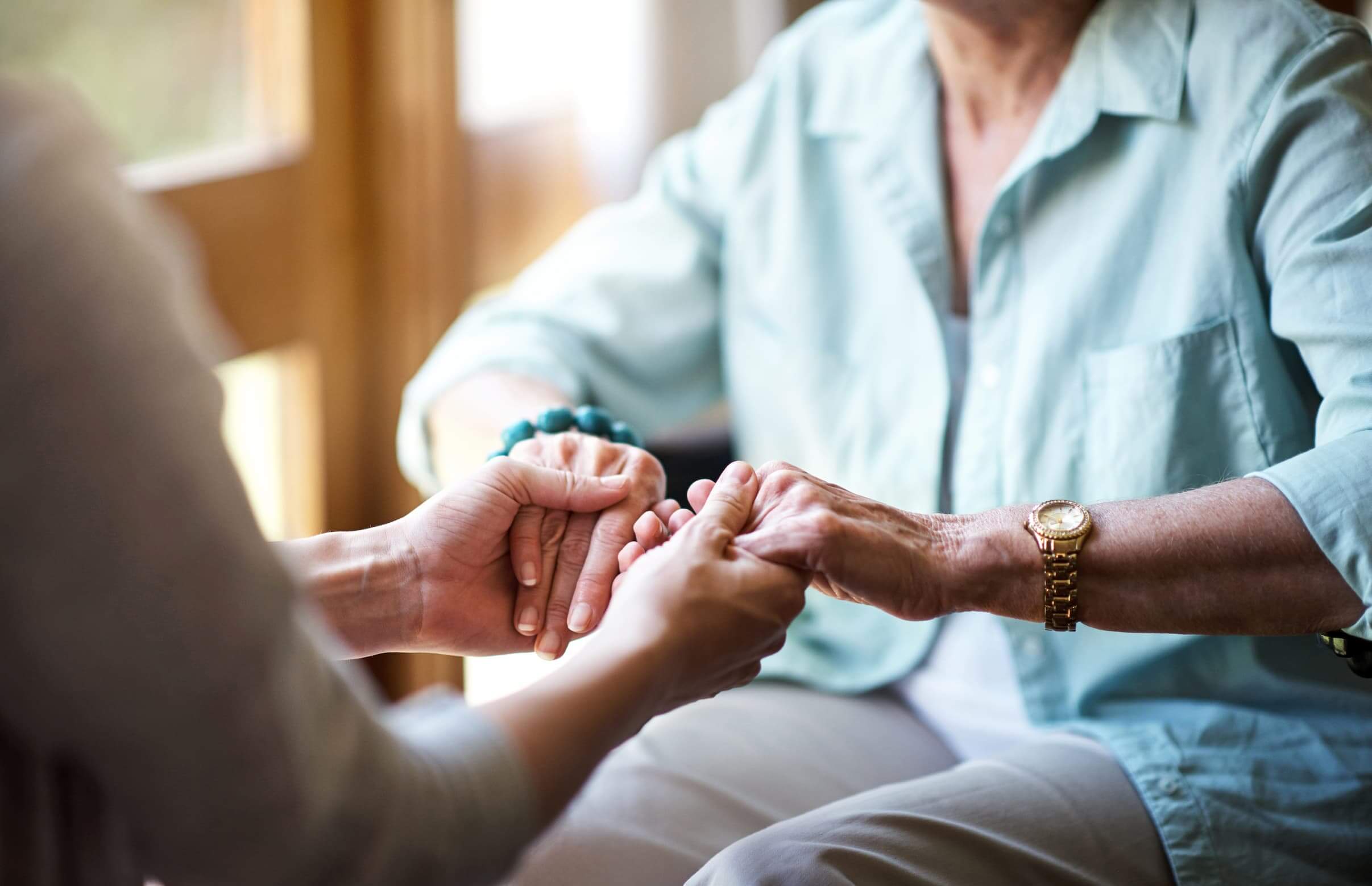 Hands of caregiver holding elderly womans hands in a warm, light-filled room.