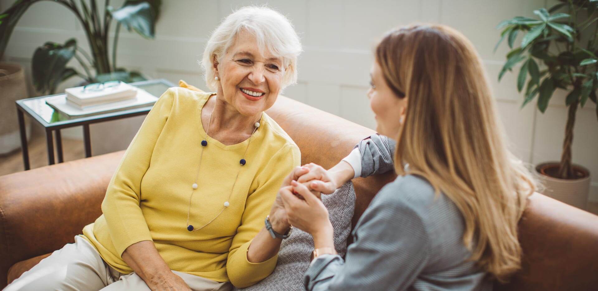 Elderly woman in yellow shirt holding hands with younger woman on a couch.