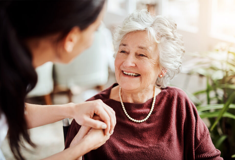 Elderly woman smiling while holding hands with a caregiver indoors.