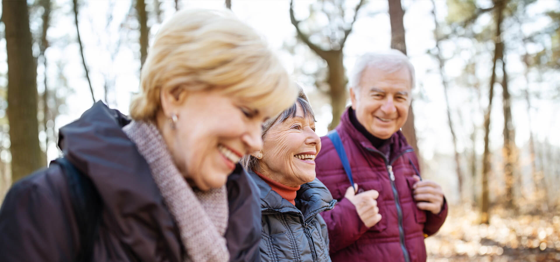 Three smiling seniors walking in a wooded area, enjoying a bright day.