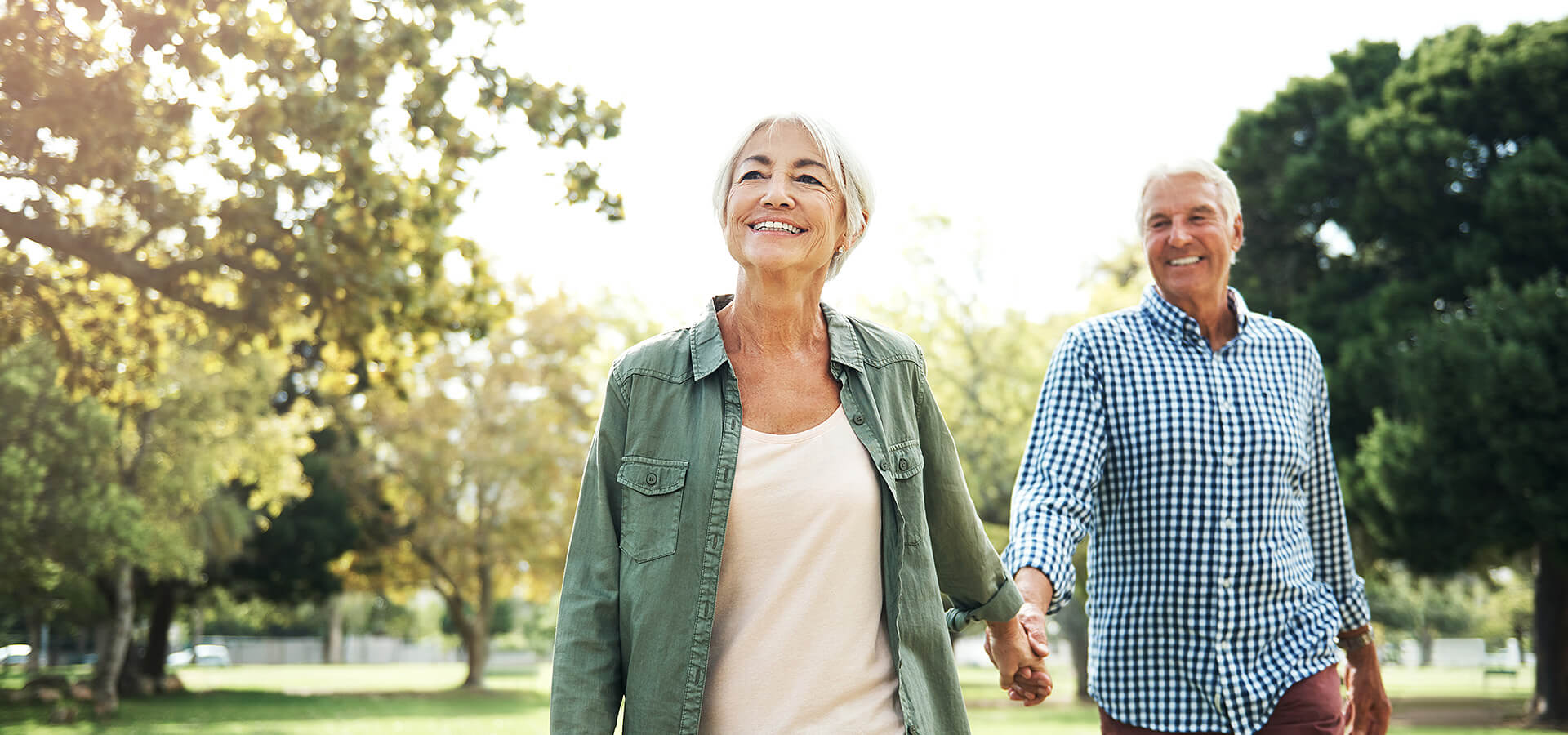 Smiling senior couple holding hands and walking in a sunny park with trees.